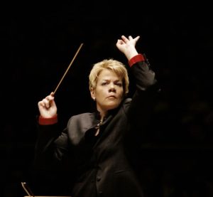 Woman holding a baton, conducting an ensemble, in front of a black background.